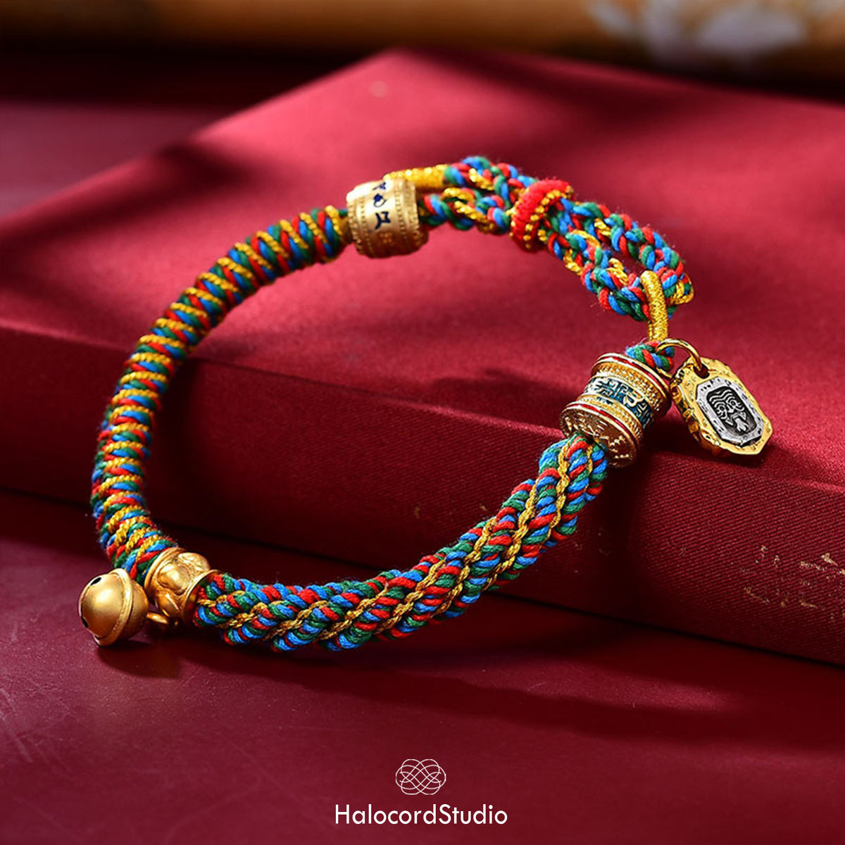 Multicolor handwoven Tibetan lucky knot cord bracelet with a golden prayer wheel bead, medallion charm, and brass bell, displayed on deep red silk