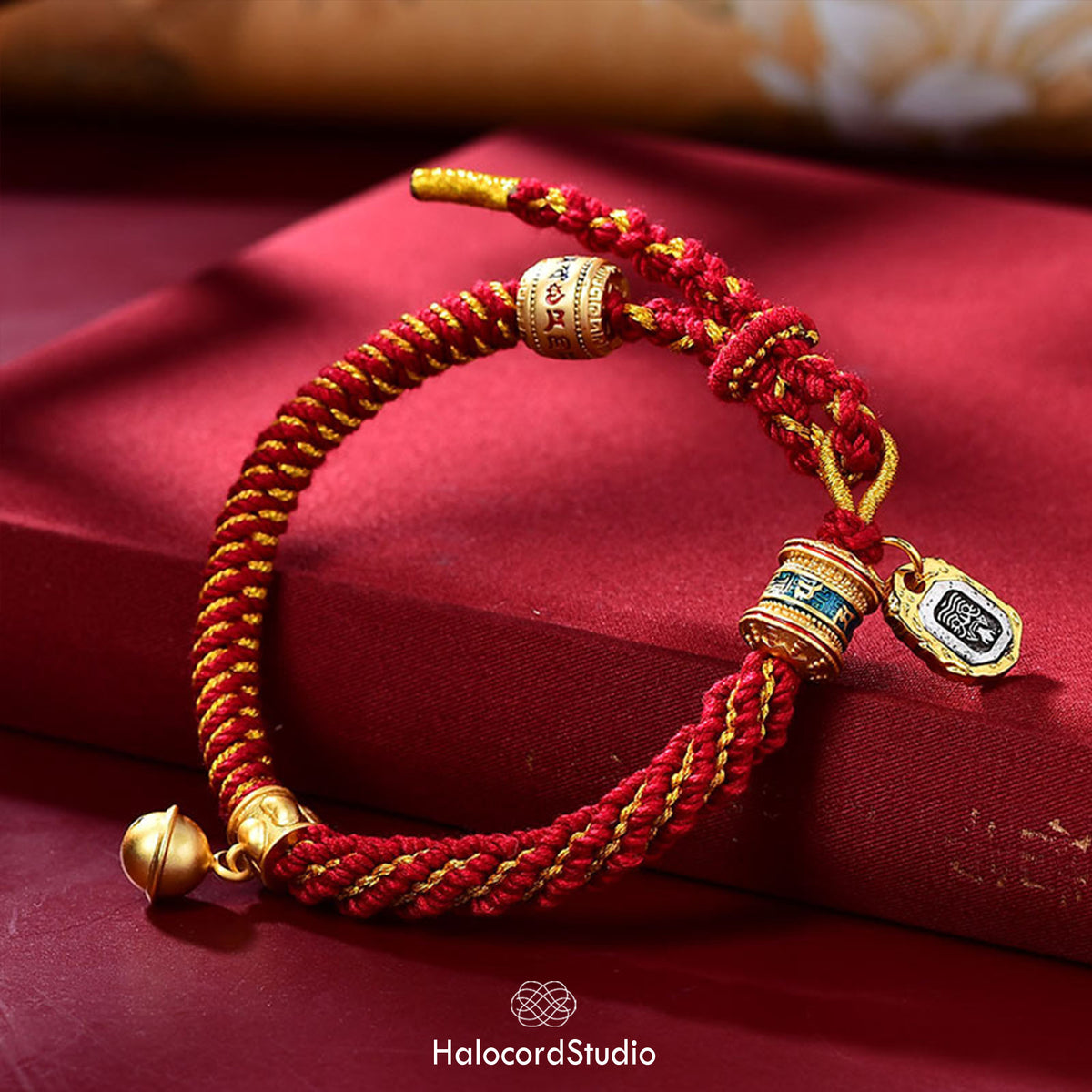 Red handwoven Tibetan lucky knot cord bracelet with a golden prayer wheel bead, medallion charm, and brass bell, displayed on red silk