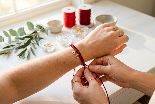 Artisan hands gently tying a red 7-knot string bracelet with a golden evil eye charm around a left wrist in warm morning light