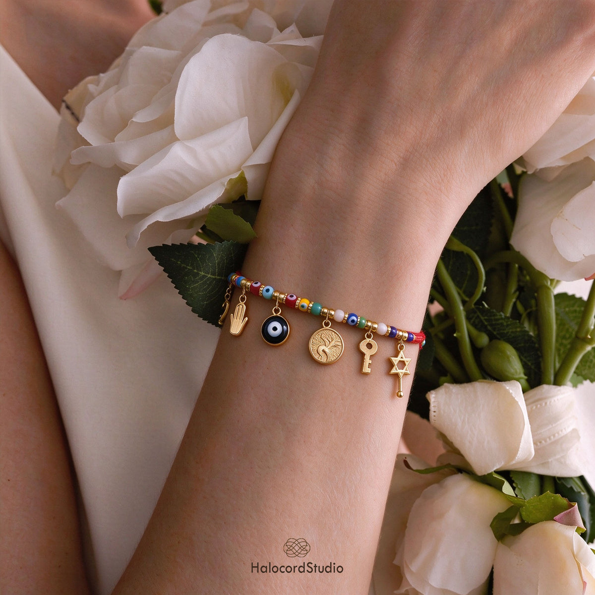 Woman wearing adjustable red cord evil eye bracelet with 8 dangling charms and colorful beads holding white roses