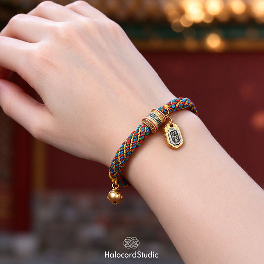 Multicolor handwoven Tibetan lucky knot bracelet with a golden prayer wheel bead, medallion charm, and brass bell, worn on a wrist in front of a temple