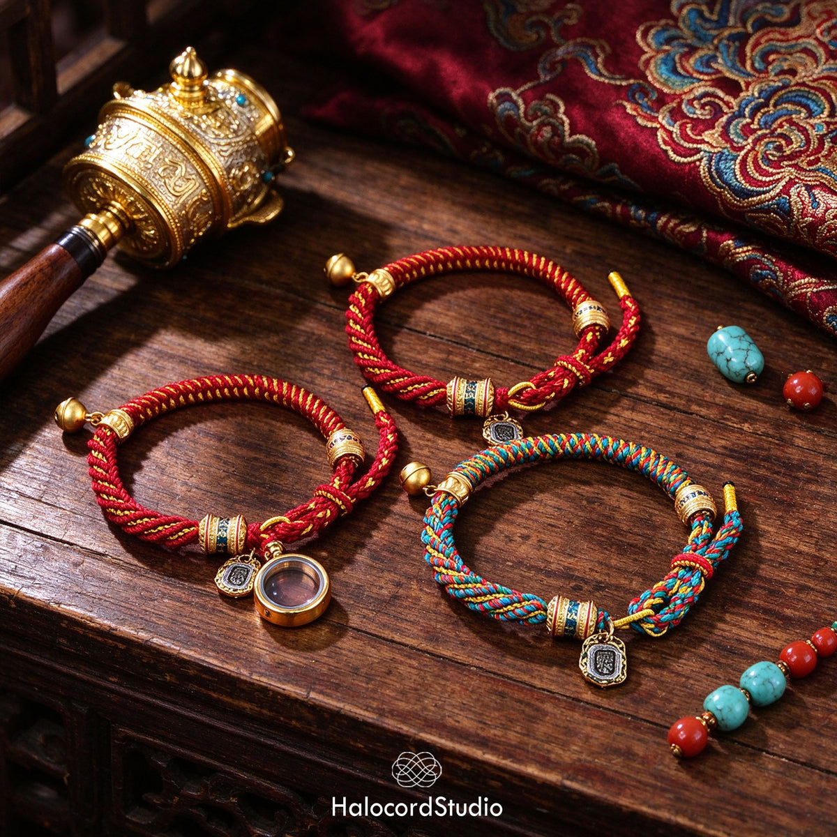 Red and multicolor handwoven Tibetan lucky knot cord bracelets with brass prayer wheel beads, bells, and a gau amulet box, styled on a wooden table with a prayer wheel and turquoise and coral beads