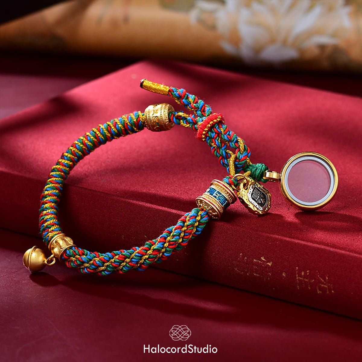 Multicolor handwoven Tibetan lucky knot bracelet with its brass gau amulet box open to reveal a red interior, displayed on red silk